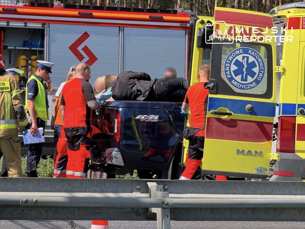 Grupa ratowników medycznych i policjantów udziela pomocy poszkodowanemu mężczyźnie w samochodzie na poboczu drogi.
