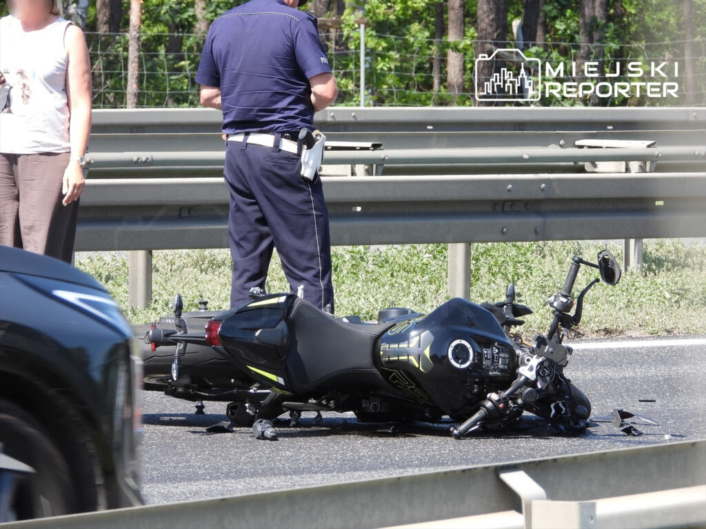Kobieta stojąca obok przewróconego motocykla na drodze, a obok niej policjant rozmawiający z nią.