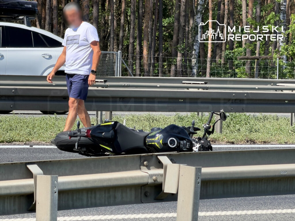 Mężczyzna w białej koszulce i szortach idzie obok przewróconego motocykla na poboczu drogi w otoczeniu drzew.