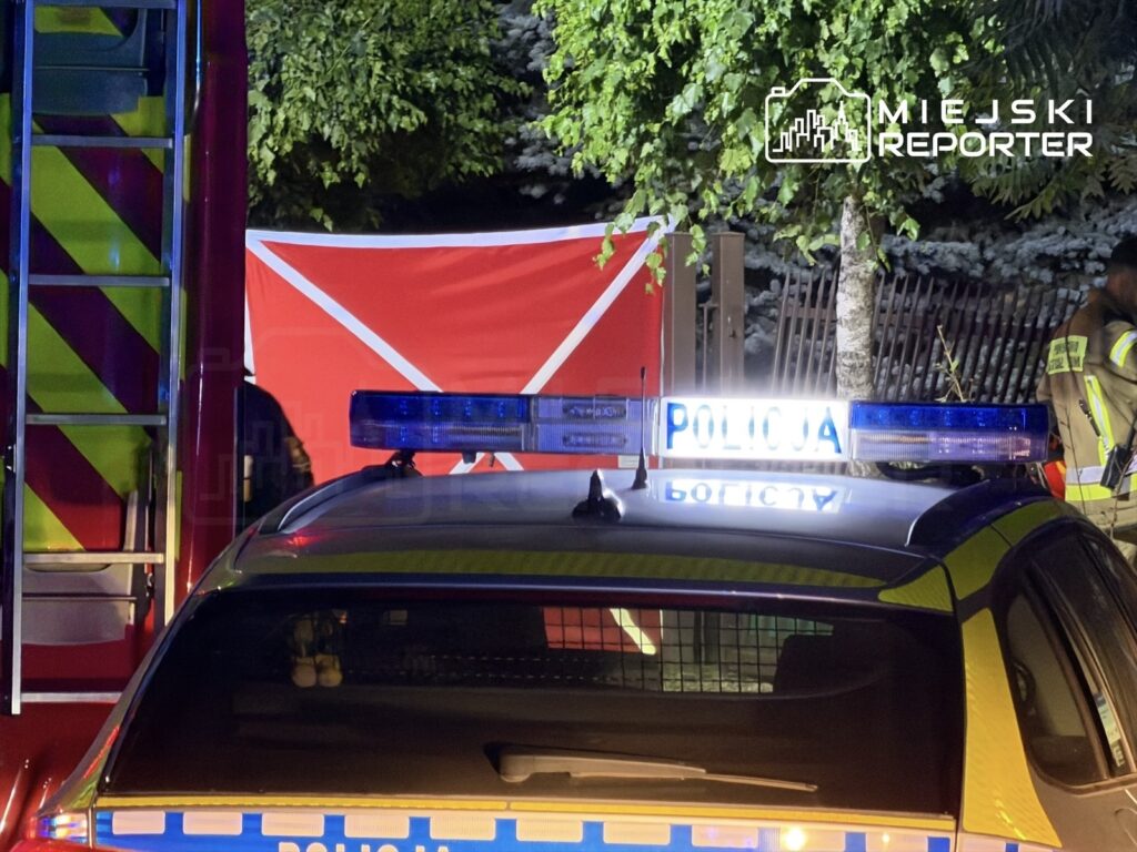 Policyjny radiowóz z włączonymi sygnałami świetlnymi stoi przed czerwonym płotem, obok drabiny strażackiej.
