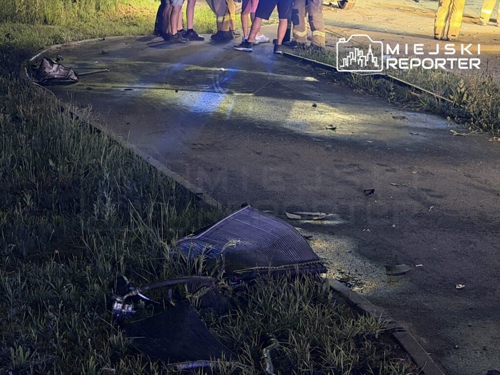 Grupa osób w odzieży roboczej omawia zdarzenie na uszkodzonej nawierzchni drogi, obok leżą fragmenty pojazdu.