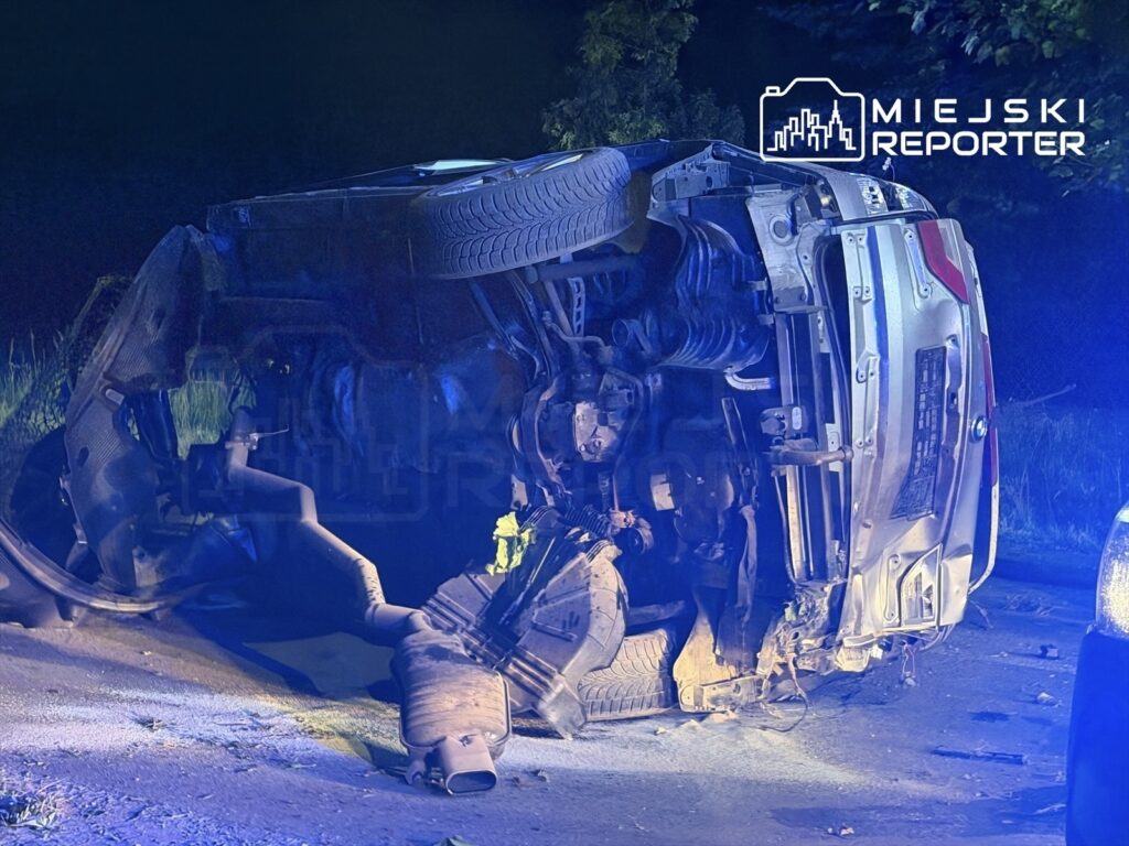 Przewrócone na bok auto leży na drodze, oświetlone niebieskimi światłami policyjnymi.
