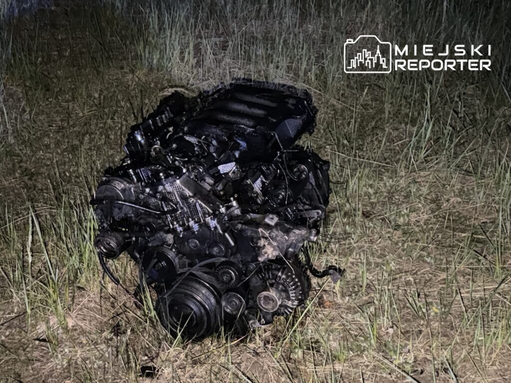 Silnik samochodowy porzucony na trawie w ciemnym otoczeniu, z widocznymi śladami użytkowania i zabrudzeniami.