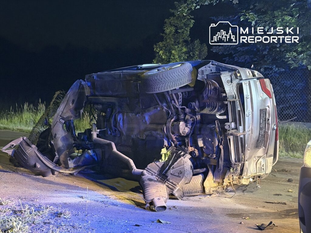 Przewrócone na dach auto leży na poboczu drogi w nocy, otoczone przez trawę i ciemność.