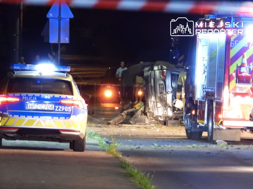 Na drodze nocą, przewrócone auto oraz pojazdy służb ratunkowych, w tym radiowóz i wóz strażacki.