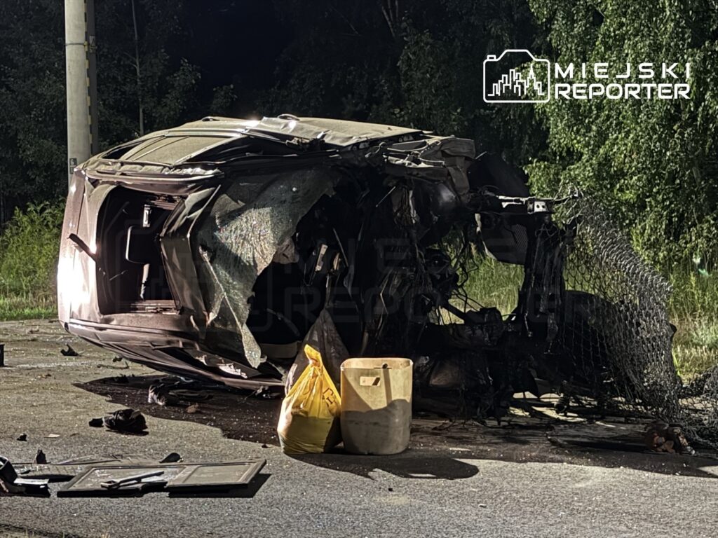 Przewrócone auto osobowe leży na boku na drodze, otoczone porozrzucanymi przedmiotami, w tle zieleń drzew.