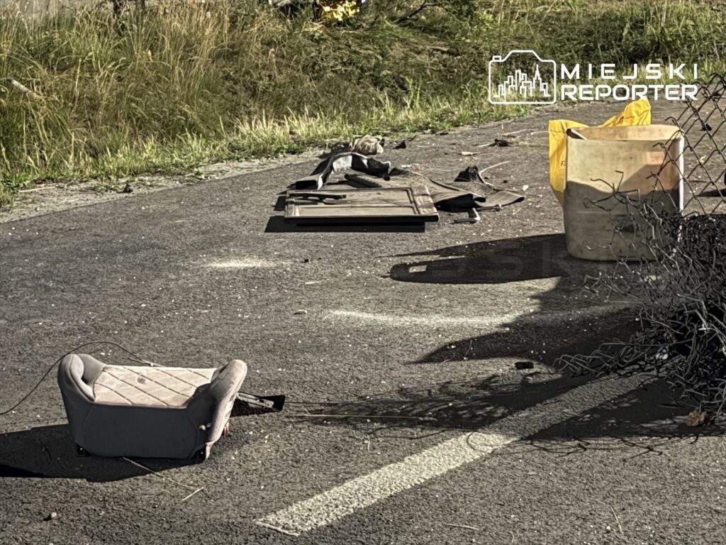 Porzucone przedmioty, w tym fotel, kawałek deski i pojemnik, leżą na asfaltowej drodze w otoczeniu trawy.
