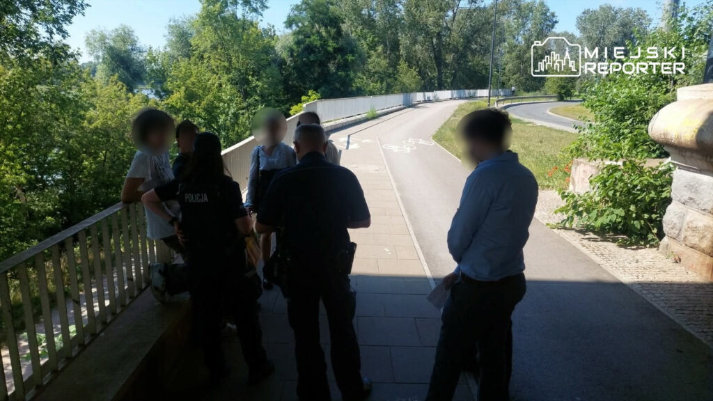Grupa młodych ludzi rozmawia z policjantami na chodniku w otoczeniu zieleni i ścieżki rowerowej.