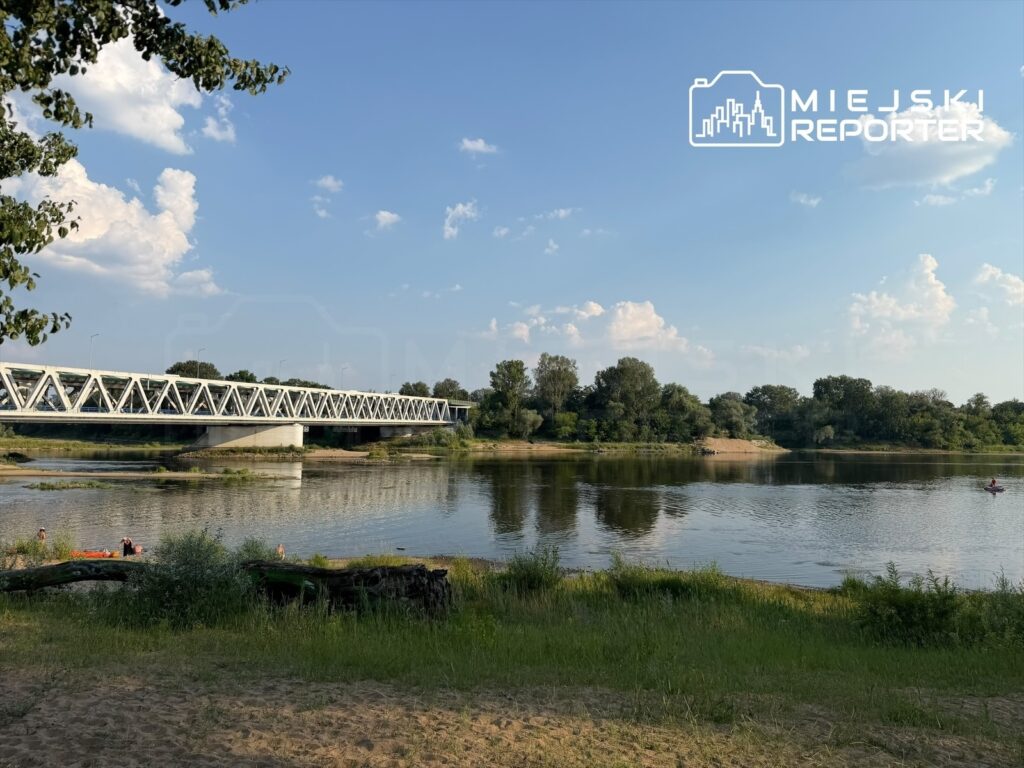 Stalowy most z przęsłami nad rzeką, w tle zieleń drzew i błękitne niebo z chmurami. Na brzegu widać ludzi wypoczywających.