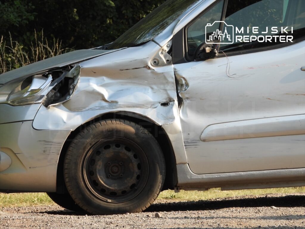 Uszkodzony samochód osobowy z wgniecioną karoserią, stojący na żwirowej drodze w otoczeniu zieleni.