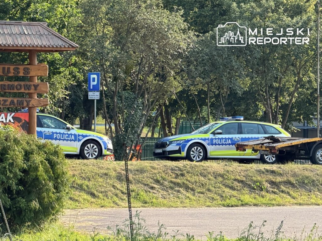Dwa radiowozy policyjne zaparkowane obok znaku informacyjnym w zielonym otoczeniu.