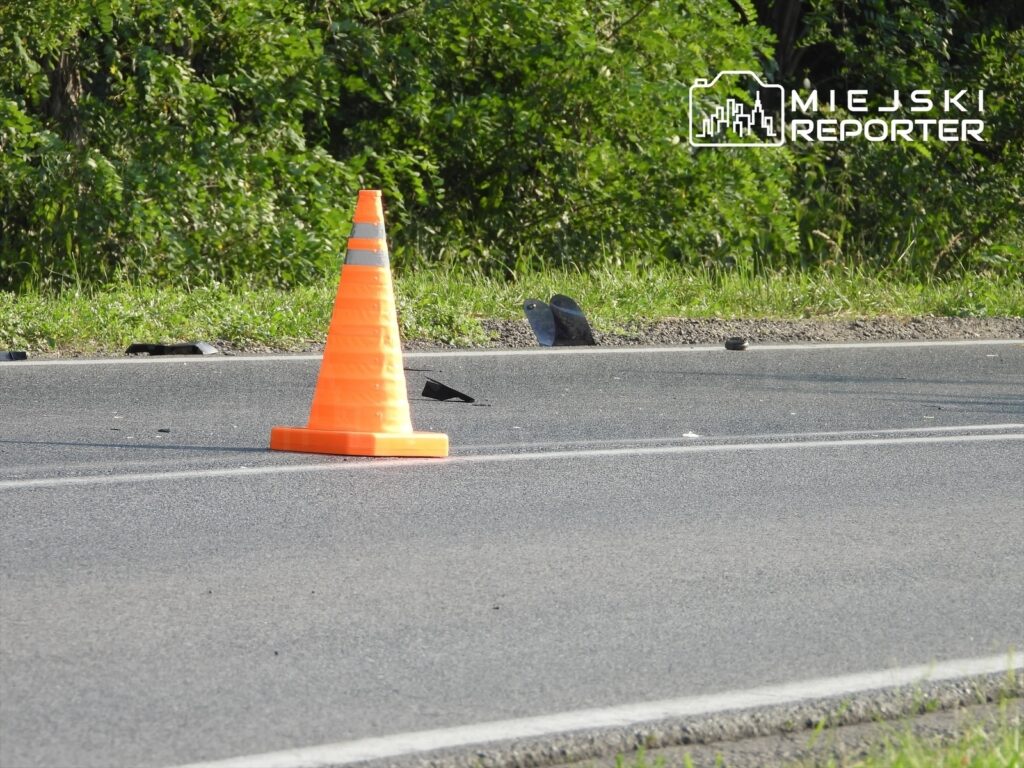 Pomarańczowy pachołek drogowy stoi na jezdni, obok leżą fragmenty sprzętu i odpadki, w tle zielona roślinność.