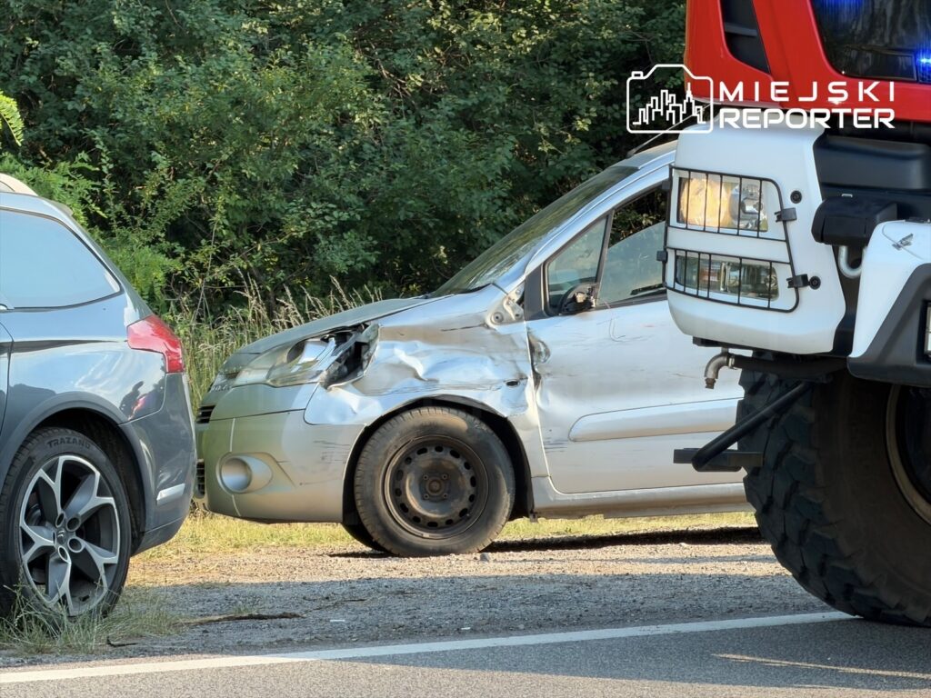 Zniszczony samochód osobowy zaparkowany na poboczu drogi, obok stoi pojazd służb ratunkowych.