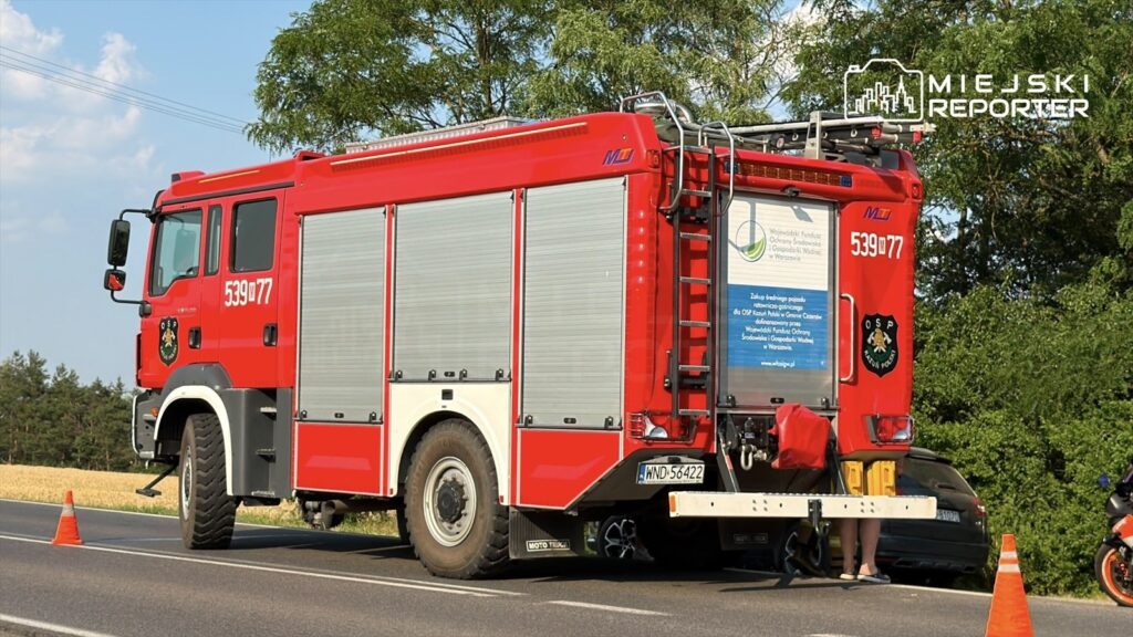 Czerwony wóz strażacki zaparkowany przy drodze, z widocznym znakiem OSP oraz osobą pracującą przy tylnej części pojazdu.