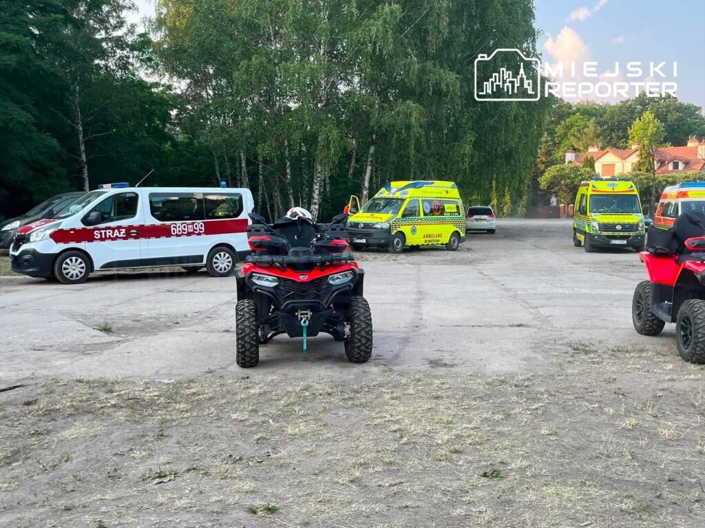 Na pierwszym planie widoczny quad, w tle kilka karetek pogotowia i pojazd straży pożarnej na parkingu otoczonym drzewami.