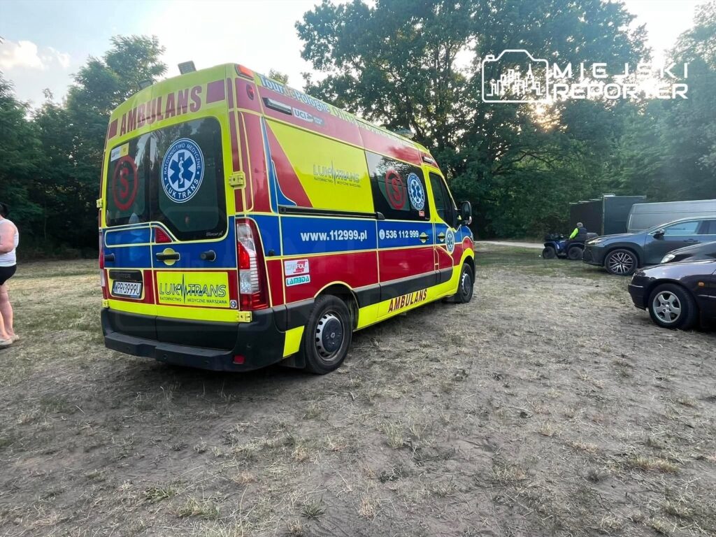 Karetka pogotowia w kolorach żółtym i czerwonym zaparkowana na trawie wśród drzew, obok kilku samochodów osobowych.