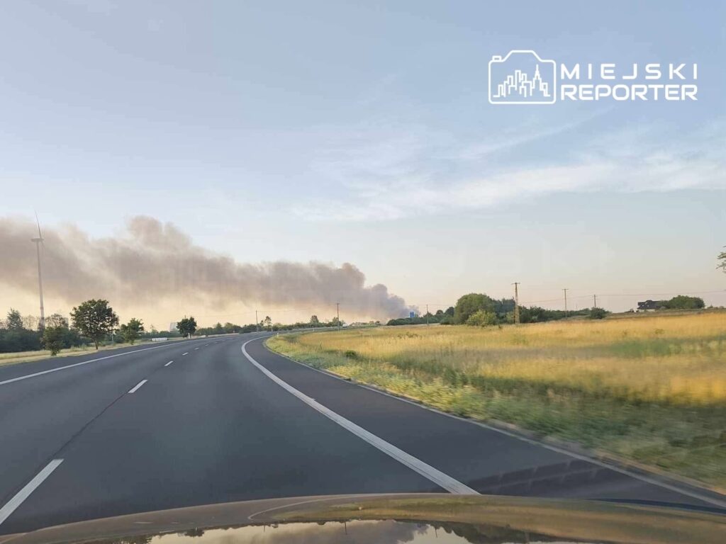 Widok na zakręt drogi, z dymem unoszącym się w tle, w otoczeniu pól i drzew.