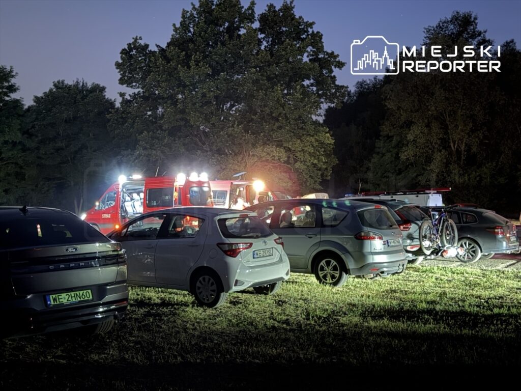 W nocy na parkingu w lesie zaparkowane samochody, w tle widać wóz strażacki z włączonymi sygnałami świetlnymi.