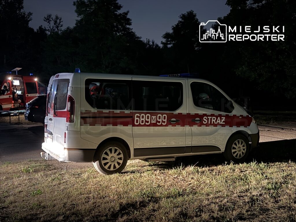 W nocy pojazd straży pożarnej zaparkowany na trawie, w tle widoczne inne pojazdy ratunkowe.