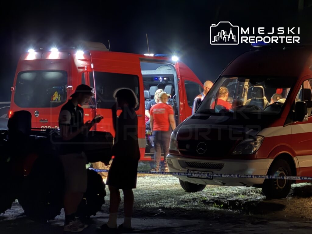 Grupa osób rozmawia w nocy obok dwóch czerwonych ambulansów, w tle widoczni ratownicy medyczni.