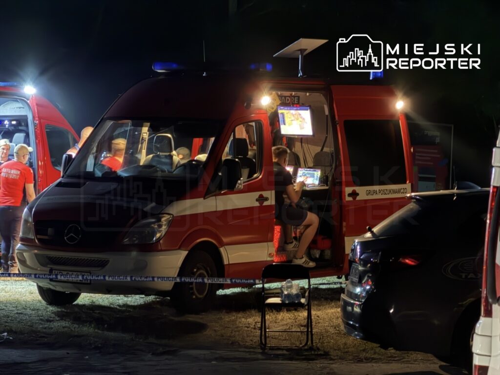 Grupa poszukiwawcza pracuje w nocy przy czerwonym ambulansie, monitorując sytuację na laptopie.