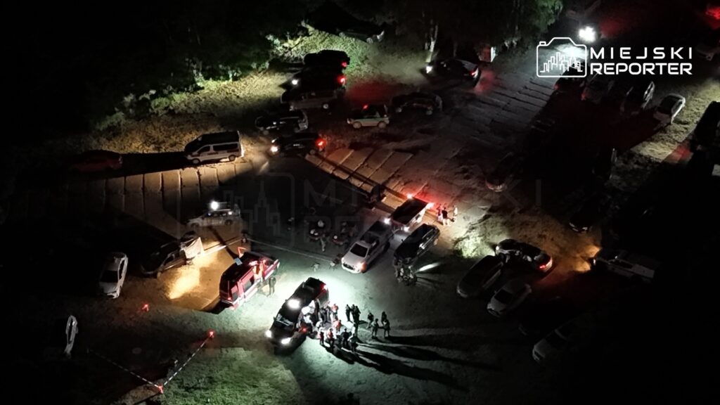 Grupa ludzi zbiera się w jasnym świetle w otoczeniu zaparkowanych samochodów na ciemnym terenie.