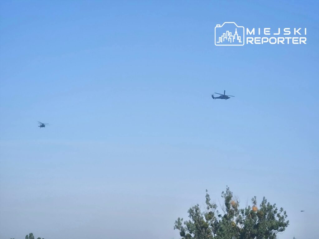 Dwa helikoptery przelatują nad zielonymi drzewami w jasnym, niebieskim niebie.