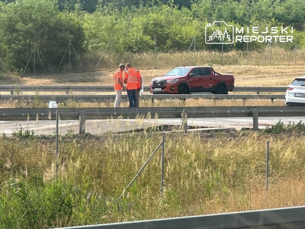 Grupa pracowników w pomarańczowych kamizelkach rozmawia obok drogi, w tle widoczny czerwony i biały samochód.