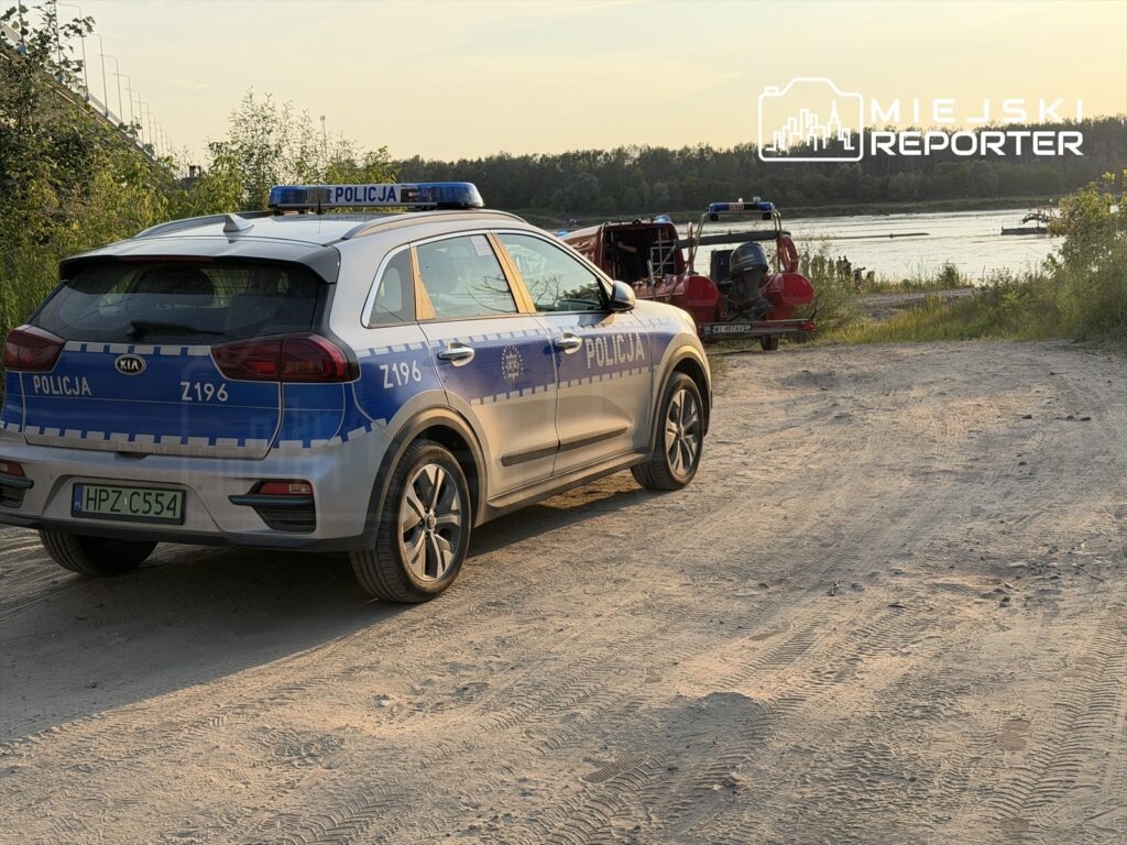 Policyjny samochód zaparkowany na piaszczystym brzegu rzeki, w tle widać łódź ratunkową z funkcjonariuszami.