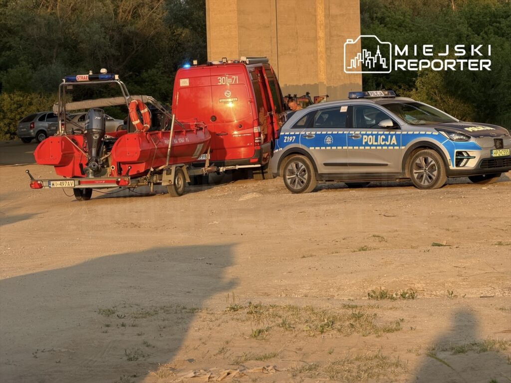 Czerwony pojazd ratunkowy z łodzią na przyczepie oraz radiowóz policyjny zaparkowane na piaszczystym terenie.