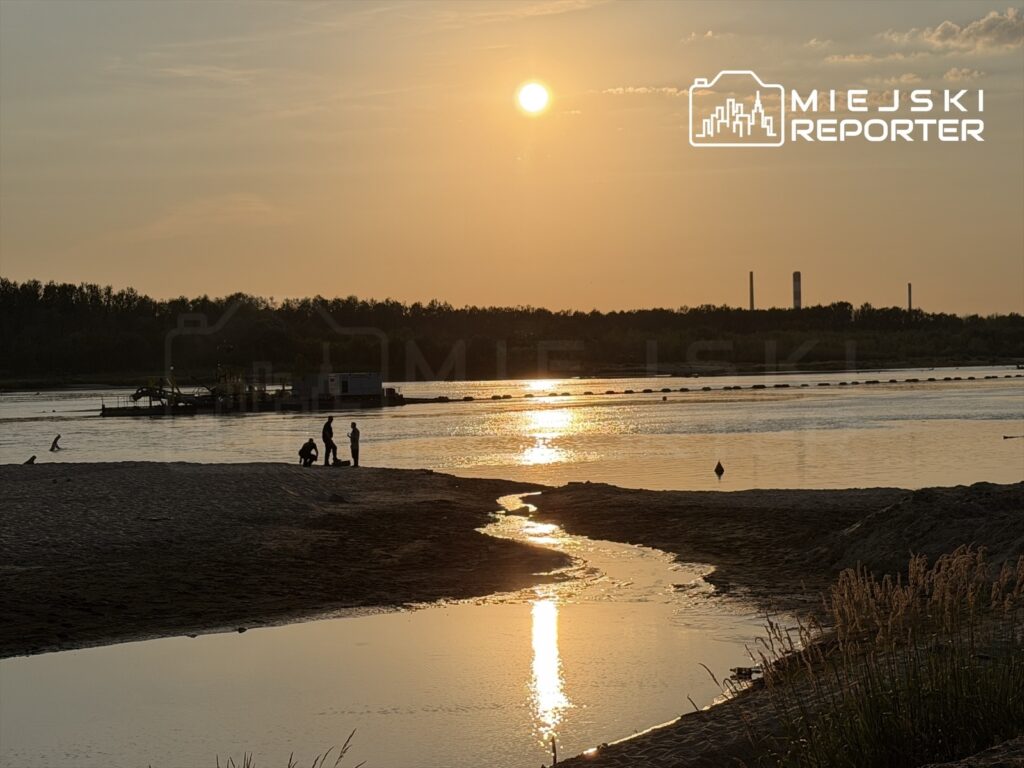 Grupa osób stoi na plaży, obserwując zachód słońca nad rzeką, w tle widoczna łódź i przemysłowe kominy.