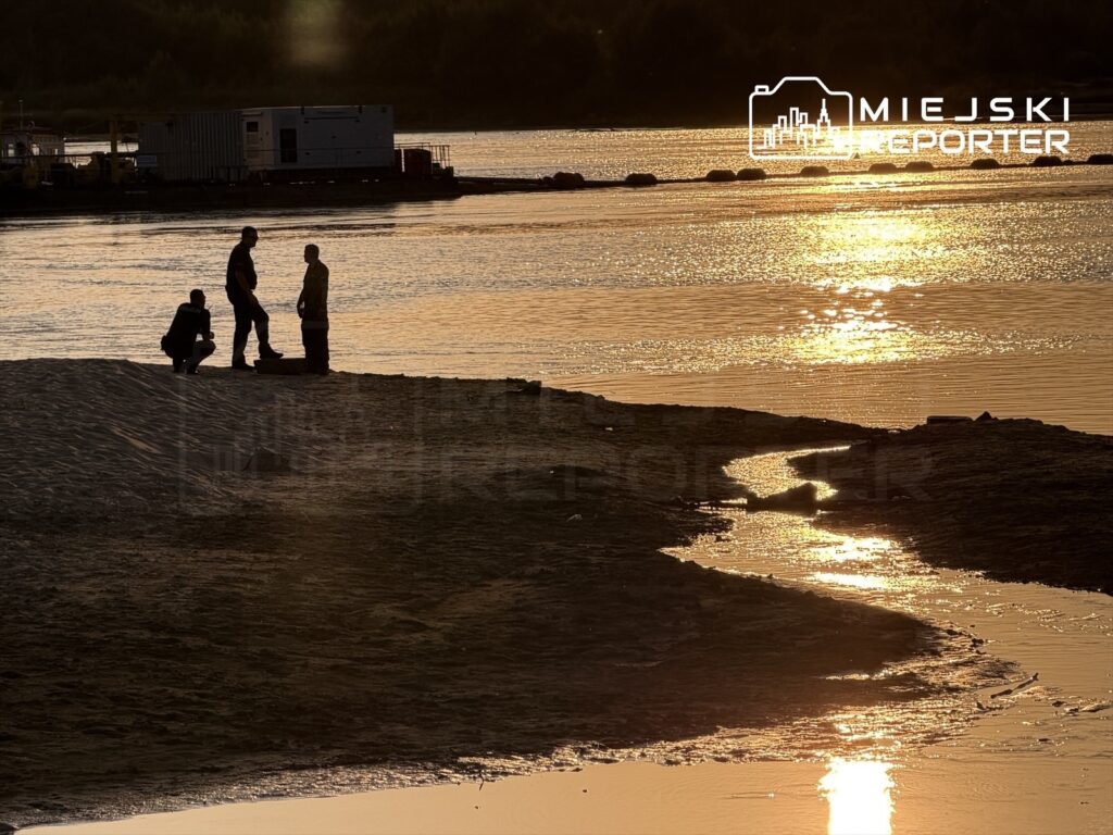 Trzech mężczyzn stoi na plaży, jeden z nich klęczy, a w tle widać odbicie słońca w wodzie.