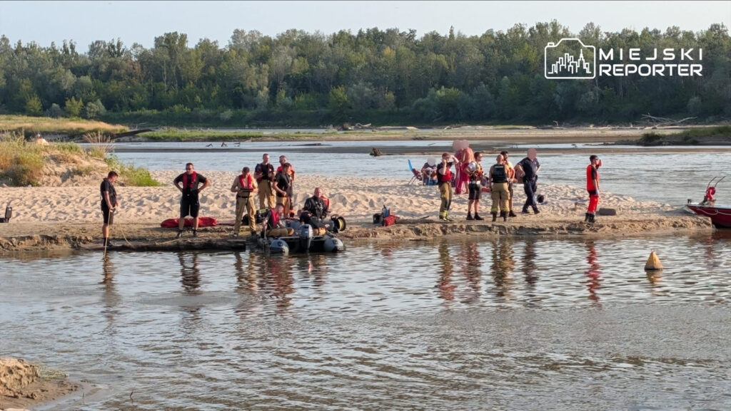 Grupa ratowników i strażaków przygotowuje sprzęt na plaży nad rzeką, w tle widoczne zarośla i wodę.