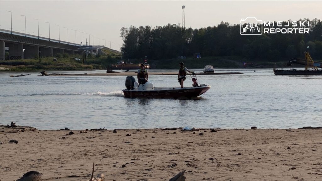 Dwóch mężczyzn w kamizelkach ratunkowych płynie łodzią motorową po rzece, w tle widoczne plaża i most.