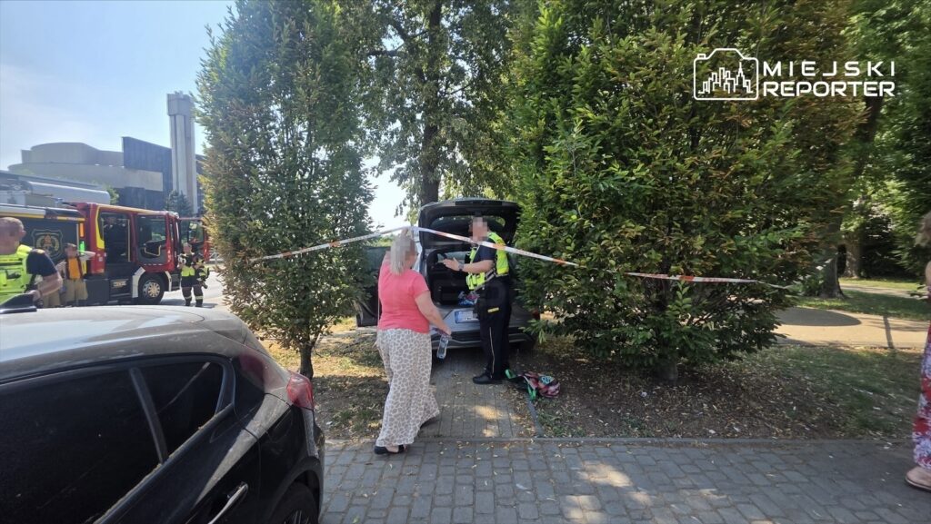 Kobieta w różowej koszulce rozmawia z policjantem obok otwartego bagażnika samochodu, w tle widać wóz strażacki.
