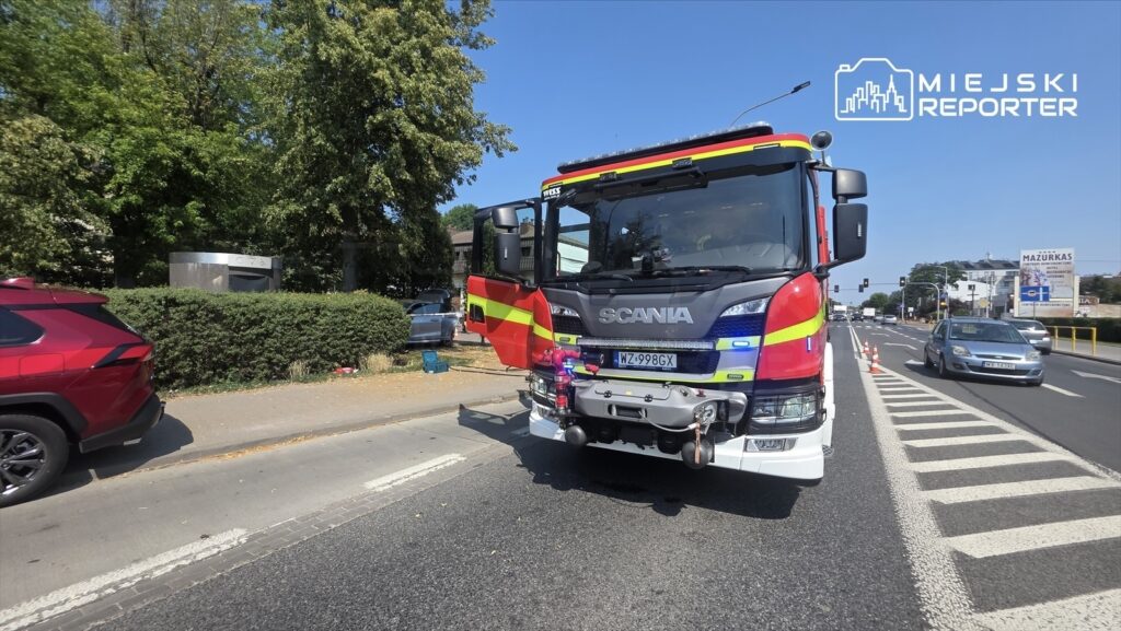Czerwony wóz strażacki marki Scania zatrzymany na ulicy, z włączonymi sygnalizatorami świetlnymi.