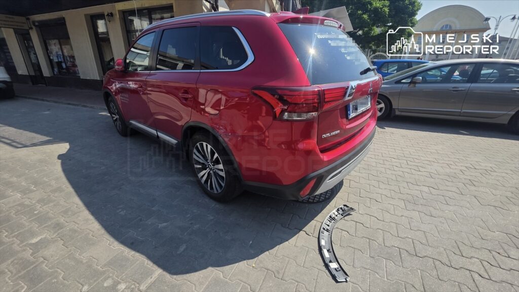 Czerwony SUV zaparkowany na ulicy, obok leży uszkodzony element karoserii.