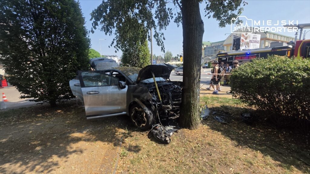 Zniszczony samochód osobowy stoi na trawie obok drzewa, z otwartą maską, w pobliżu przejeżdżających pojazdów i przechodniów.