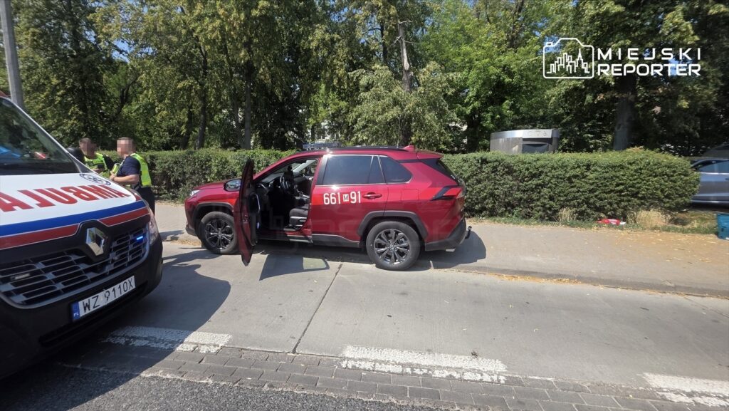Na ulicy stoi czerwony SUV z otwartymi drzwiami, obok dwóch policjantów rozmawia z osobą w mundurze.