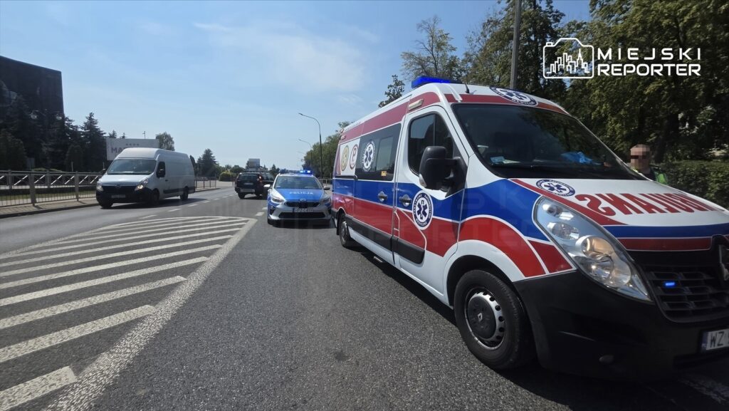 Karetka pogotowia z włączonymi sygnałami świetlnymi zatrzymana na jezdni, obok radiowóz policyjny.