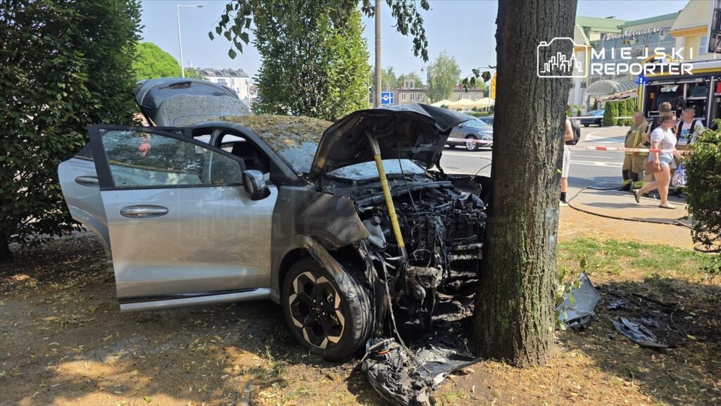 Uszkodzony samochód osobowy stoi przy drzewie, z otwartą maską i uszkodzoną przednią częścią, w otoczeniu przechodniów.