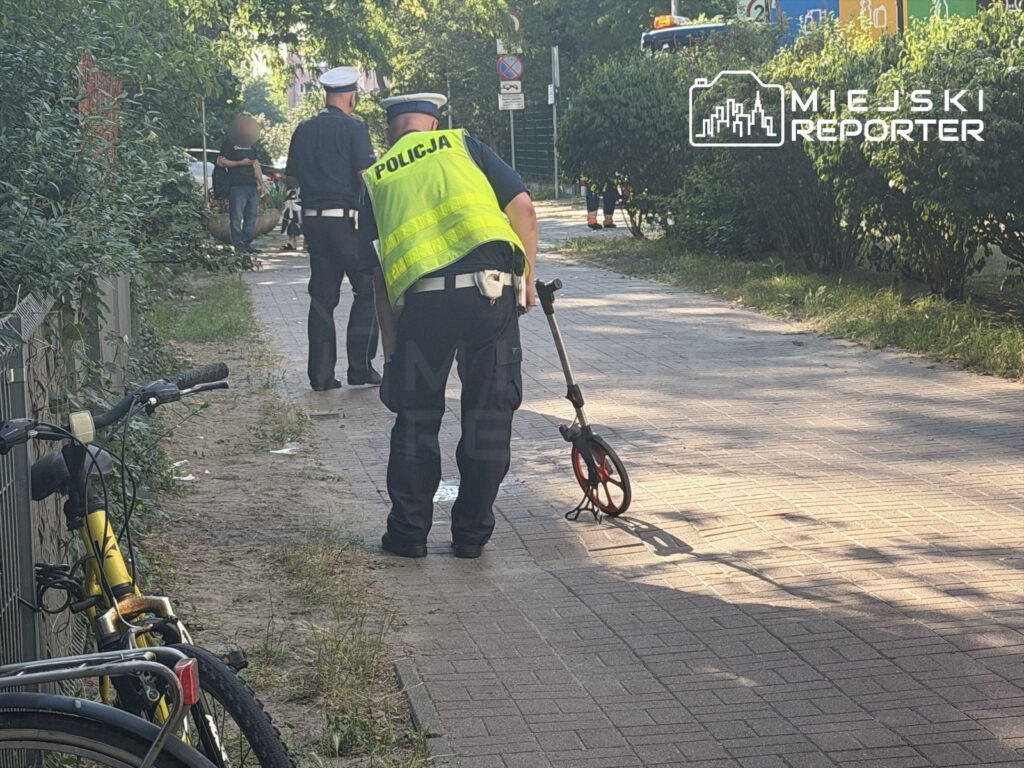 Dwaj policjanci w mundurach, jeden mierzy odległość specjalnym urządzeniem na chodniku, drugi obserwuje otoczenie.
