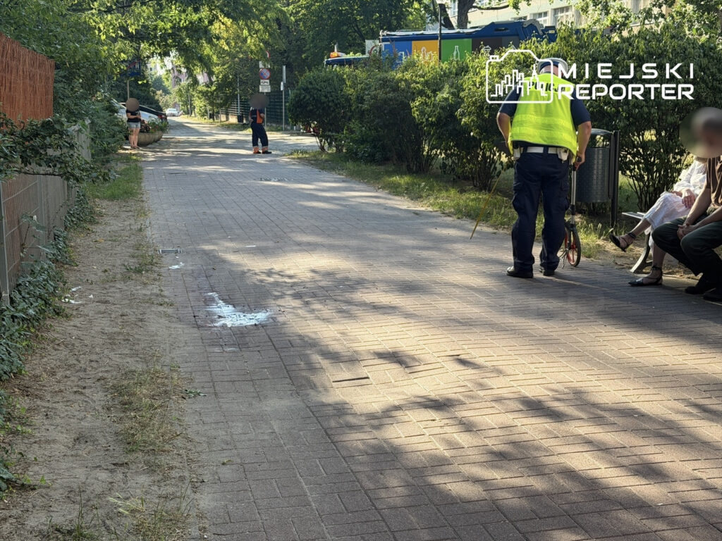 Mężczyzna w kamizelce odblaskowej rozmawia z dwoma osobami siedzącymi na ławce w parku. W tle widoczna ulica.