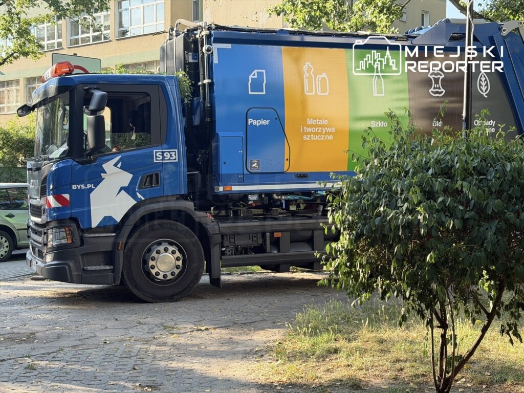Śmieciarka z kolorowymi oznaczeniami na boku parkuje na osiedlowym parkingu, otoczona zielenią drzew i krzewów.