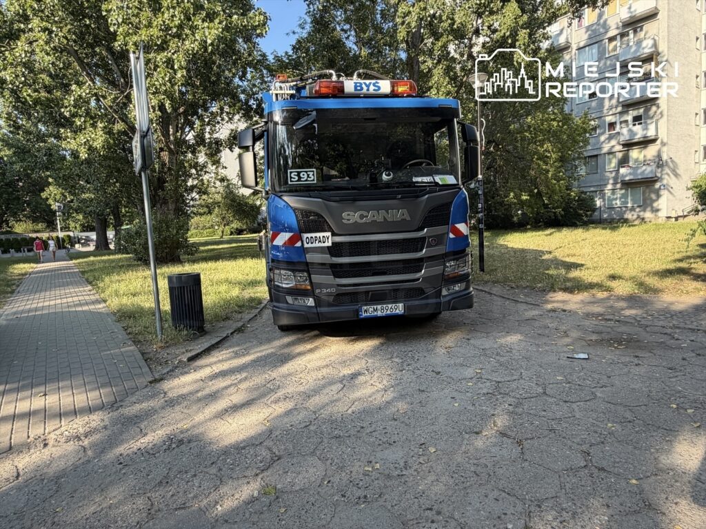 Samochód ciężarowy marki Scania z napisem "ODPADY" zatrzymany na chodniku w parku, obok śmietnika.