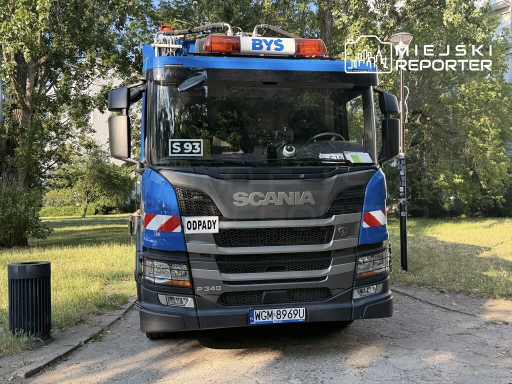 Samochód ciężarowy Scania z oznaczeniem "ODPADY" stoi na chodniku w parku, otoczony zielenią.