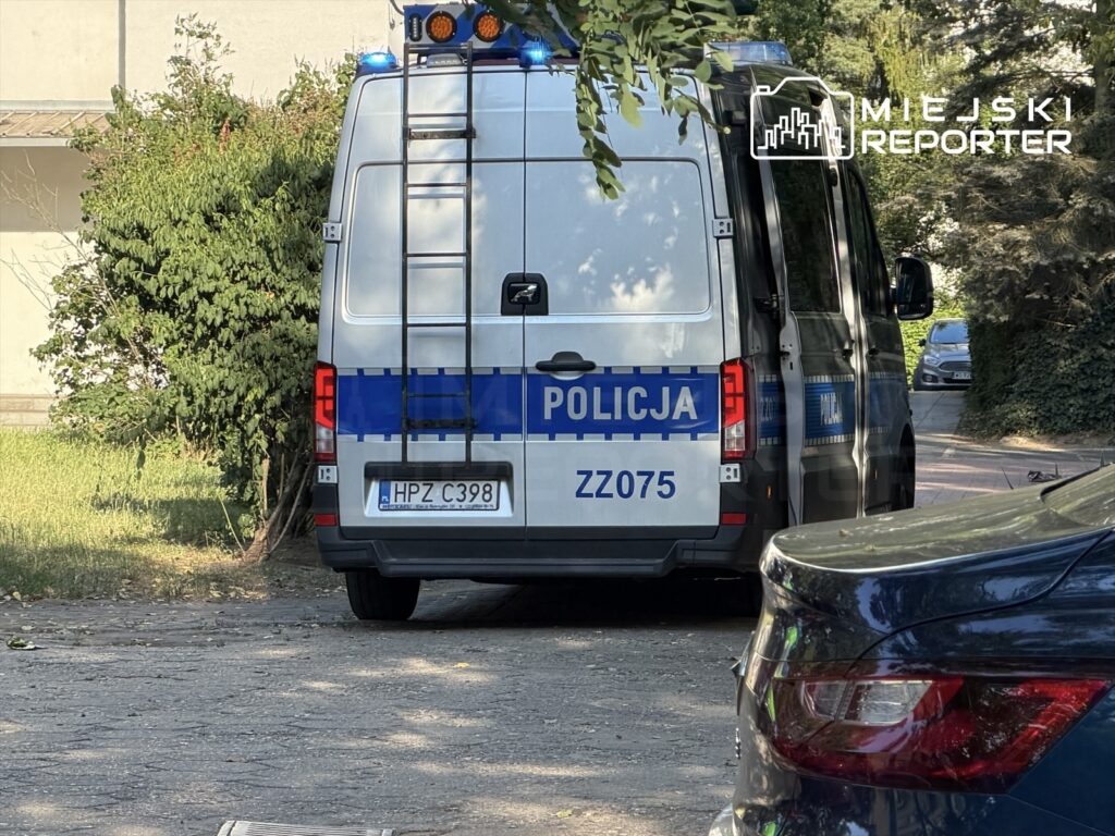 Funkcjonariusze policji w radiowozie zaparkowanym przy zadrzewionej ulicy.