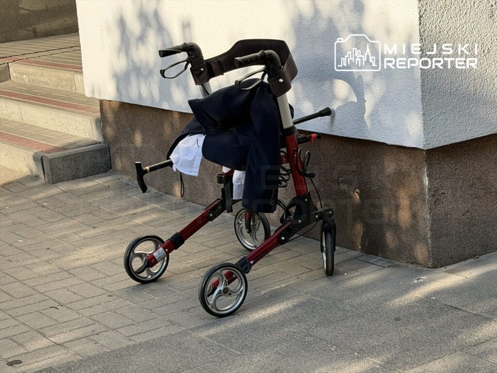 Czerwony balkonik rehabilitacyjny oparty o ścianę, obok leży ciemna kurtka. W tle widoczne schody.