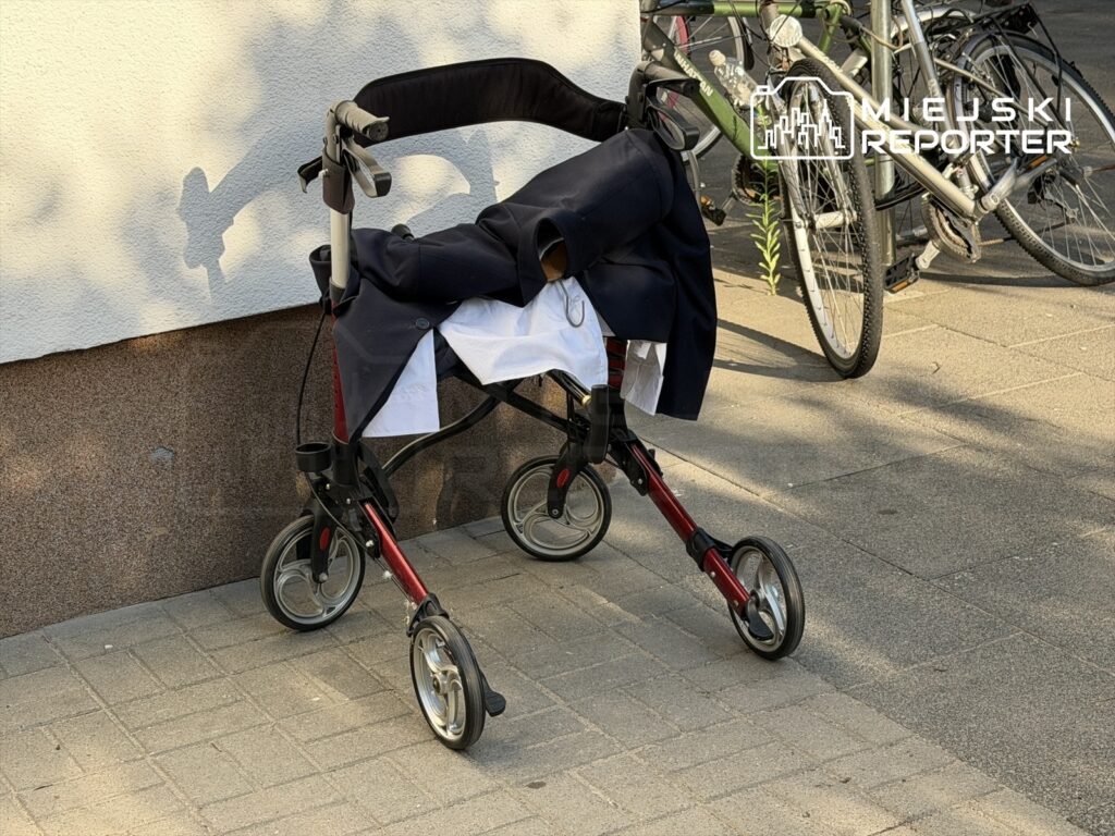 Wózek inwalidzki z ciemnym płaszczem leżącym na siedzisku, ustawiony na chodniku obok rowerów.