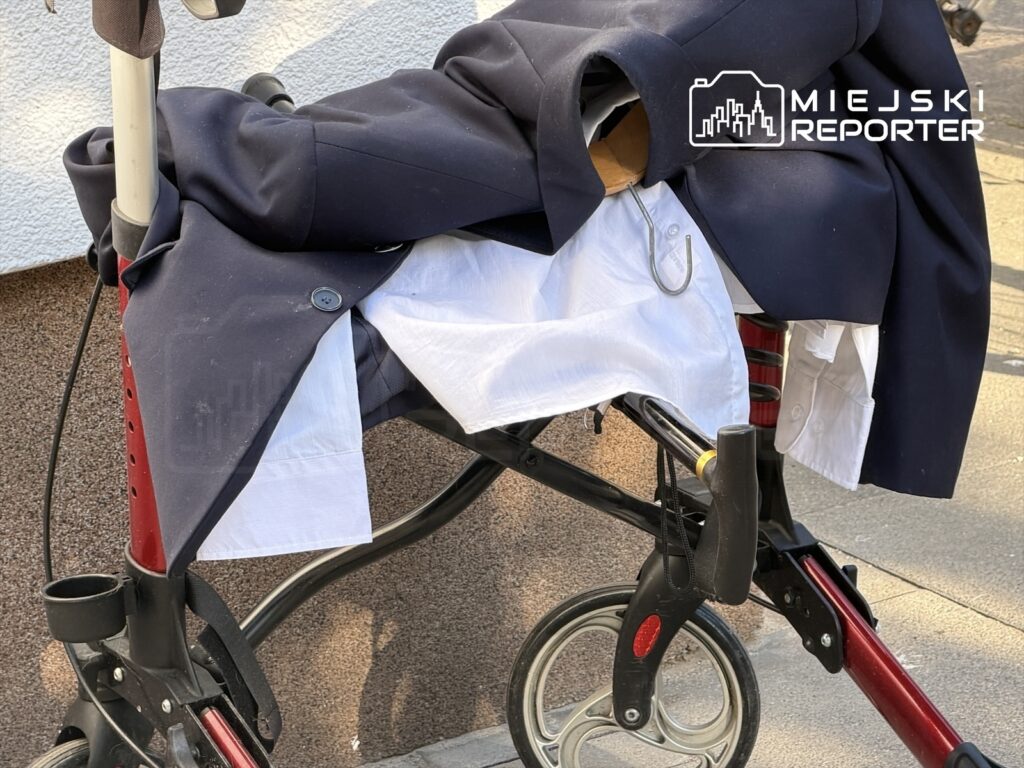 Wózek inwalidzki z nałożoną marynarką i białą koszulą, stojący na chodniku obok ściany budynku.